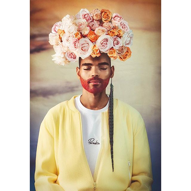 A man with a floral headpiece and red beard stands serenely in a meditative portrait.