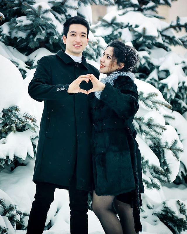 A couple makes a heart with their hands in a snowy park. They are dressed in winter coats and smiling.