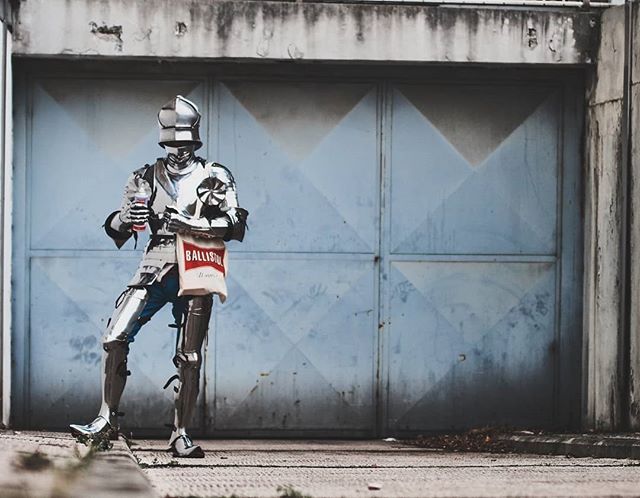 A knight in full armor humorously holds a soda and shopping bag in an urban setting, creating an amusing editorial image.