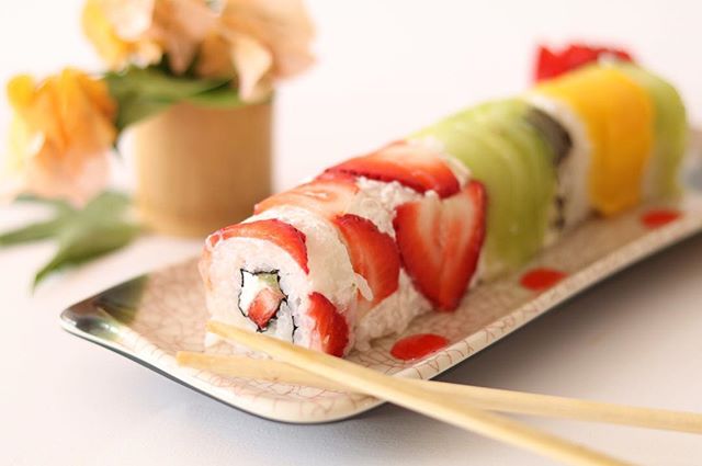 A colorful fruit sushi roll with strawberry, kiwi, and mango on a decorative plate with chopsticks.