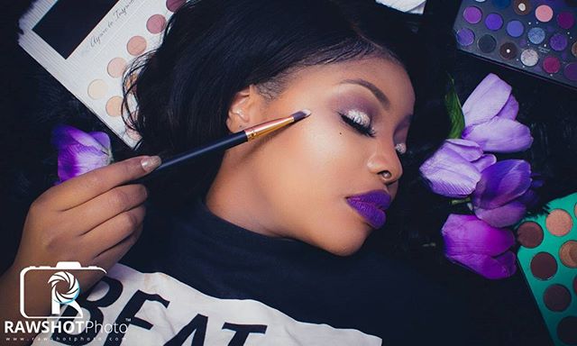 A woman with creative makeup is lying down with makeup palettes and flowers.