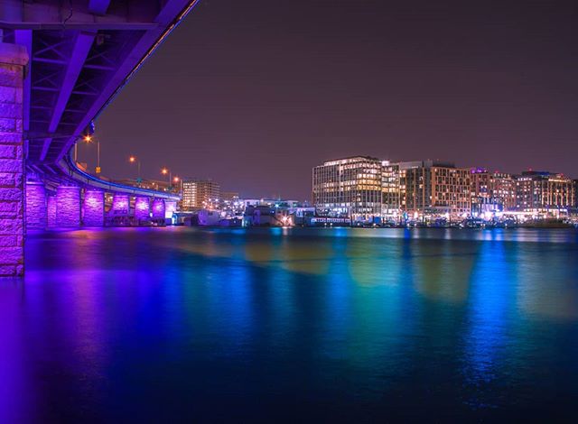 A nighttime view of a city skyline with a bridge illuminated in purple light over calm waters reflecting the city lights.