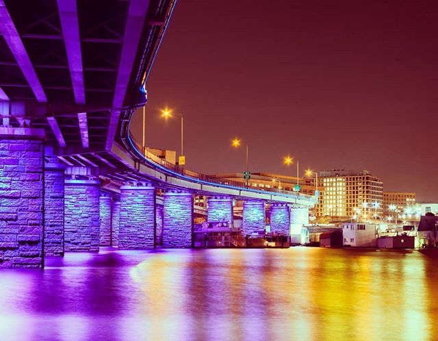 A bridge illuminated at night, spans across the water in an urban cityscape, reflecting the lights.