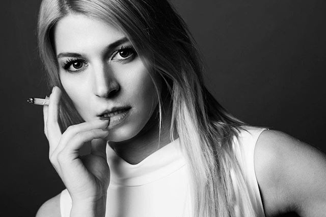 A black and white portrait of a blonde woman with a cigarette, looking confidently at the camera.